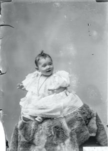 A baby sitting up on a raised platform wearing a christening dress and necklace.