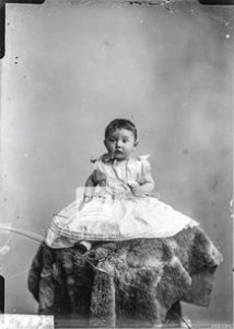 A baby sitting up on a raised platform wearing a christening dress and necklace.